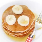 Vegane Bananenpfannkuchen auf einem Teller, dekoriert mit frischen Bananen und Sirup.