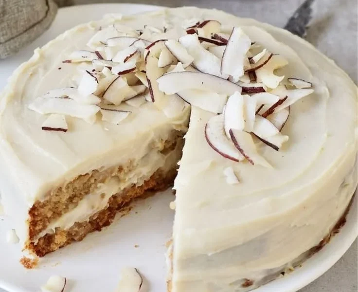Veganer Kokosnusskuchen auf einem Holzbrett mit Kokosraspeln dekoriert.