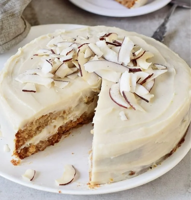 Veganer Kokosnusskuchen auf einem Holzbrett mit Kokosraspeln dekoriert.